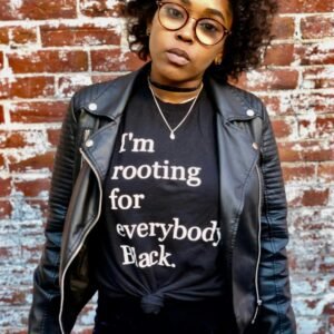 Confident Black woman in a leather jacket and statement tee against a brick wall.